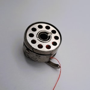 May include: A silver bobbin with a red thread attached. The bobbin has a circular design with multiple holes.