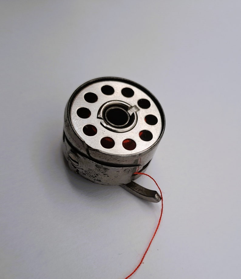 May include: A silver bobbin with a red thread attached. The bobbin has a circular design with multiple holes.