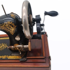 May include: An antique black sewing machine with gold floral detailing. The machine features a large silver flywheel, a black handle, and sits on a wooden base. The design is ornate and classic, showcasing craftsmanship.