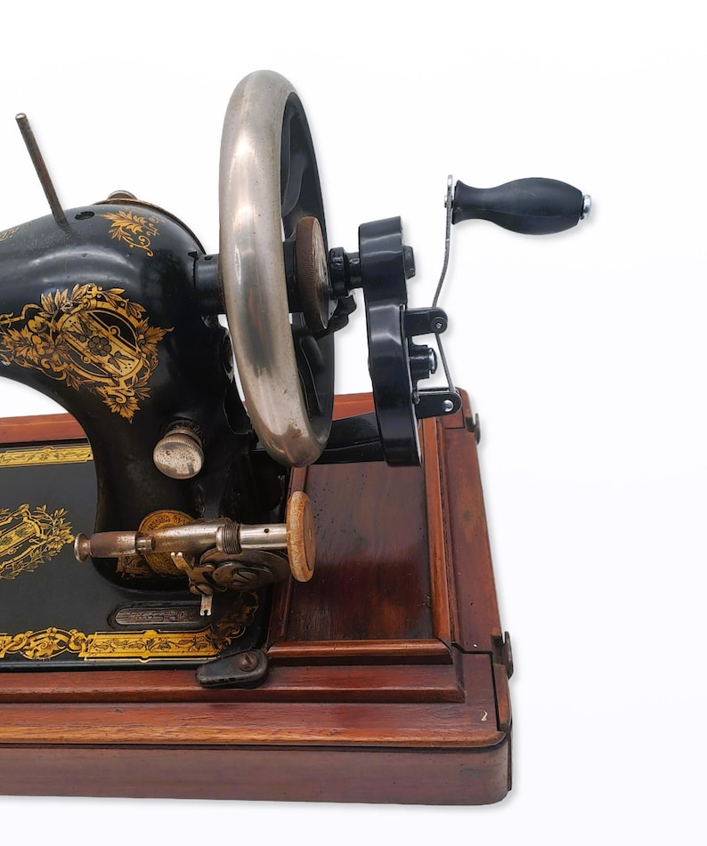 May include: An antique black sewing machine with gold floral detailing. The machine features a large silver flywheel, a black handle, and sits on a wooden base. The design is ornate and classic, showcasing craftsmanship.