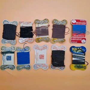 May include: A collection of vintage thread spools in various colors, including black, brown, and blue. The spools have decorative labels with text and are arranged on a peach-colored surface. Some labels read "MAAS" and "WOL".