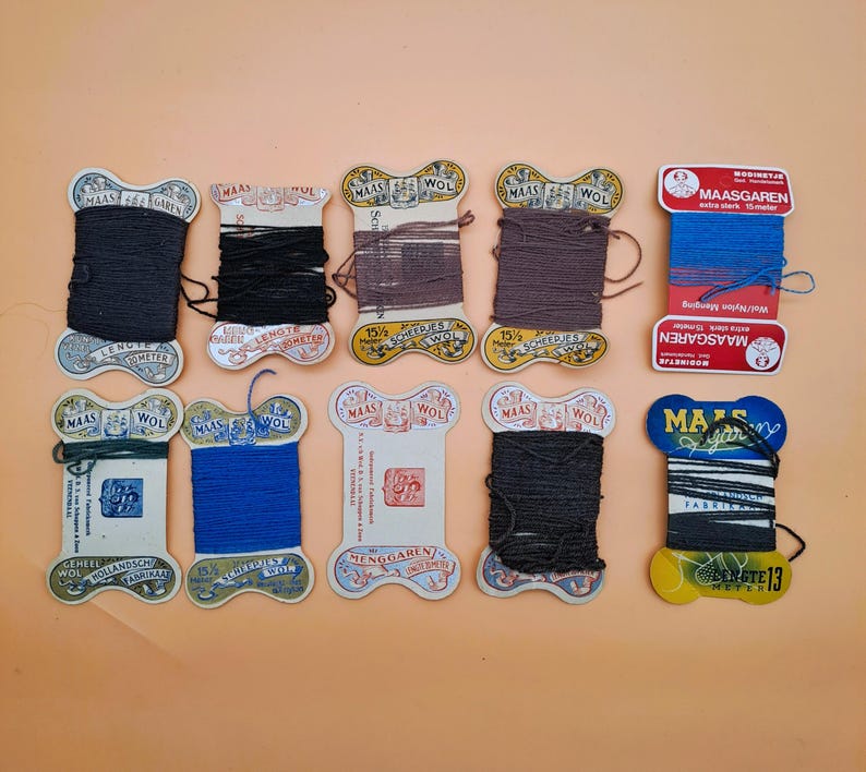May include: A collection of vintage thread spools in various colors, including black, brown, and blue. The spools have decorative labels with text and are arranged on a peach-colored surface. Some labels read "MAAS" and "WOL".
