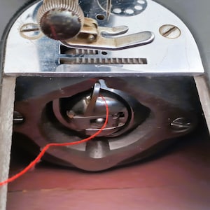 May include: Close-up of a sewing machine bobbin with red thread wound around it. The bobbin is in a metal casing and is visible through a clear plastic window.