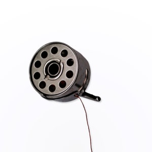 May include: A silver metal bobbin with a red thread attached. The bobbin has a circular shape with multiple holes around the center.