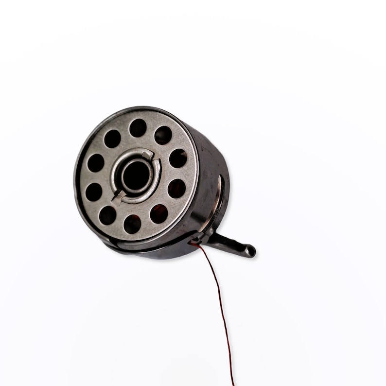 May include: A silver metal bobbin with a red thread attached. The bobbin has a circular shape with multiple holes around the center.