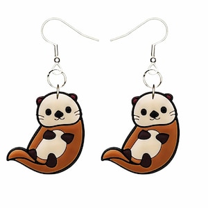 Pode incluir: Um par de brincos em forma de lontra. Cada brinco apresenta um desenho de lontra de desenho animado com rosto e barriga creme, corpo castanho e detalhes em castanho escuro. Os brincos têm fechos de gancho prateados.