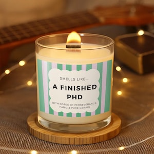 Puede incluir: Una vela encendida en un tarro de cristal transparente con una etiqueta que dice "Smells like... A Finished PHD". La etiqueta tiene un fondo a rayas verdes menta y blancas. La vela está sobre un posavasos redondo de madera.