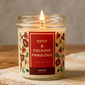Peut inclure: Une bougie en cire de soja allumée dans un bocal en verre. L'étiquette indique "FIFTY & FUCKING FABULOUS" en blanc sur fond rouge, avec des images de lèvres, de fraises et de cocktails. La bougie est posée sur une surface en bois.