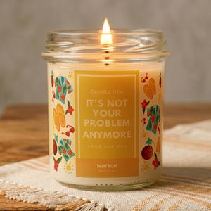 May include: A lit candle in a clear glass jar with a yellow label featuring the text 'Smells like IT'S NOT YOUR PROBLEM ANYMORE' and colourful illustrations of tropical shirts, flip-flops, sunglasses, and flowers on a white background.