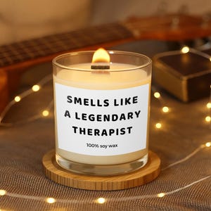 Può includere: Una candela accesa in un barattolo di vetro trasparente con la scritta "Smells Like A Legendary Therapist" su un'etichetta bianca. La candela è appoggiata su un sottobicchiere rotondo di legno. L'etichetta riporta anche "100% soy wax".