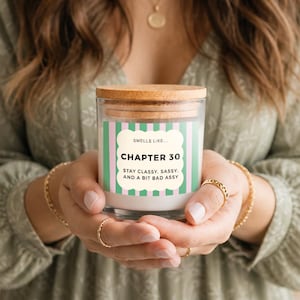 Op de afbeelding: Een glazen kaarsenpot met een houten deksel. Het etiket luidt "Chapter 30" met de tekst "Stay classy, sassy, and a bit bad assy." Het etiket heeft een groen en wit gestreept ontwerp. De kaars wordt vastgehouden voor een persoon die een groene jurk draagt.