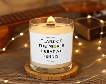 Puede incluir: Una vela encendida en un tarro de cristal transparente con una mecha de madera, sobre un posavasos redondo de madera. La etiqueta dice "SMELLS LIKE... TEARS OF THE PEOPLE I BEAT AT TENNIS". La vela está rodeada de luces de cadena.