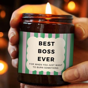 Op de afbeelding: Een brandende kaars in een bruine glazen pot met een groen en roze gestreept etiket. Op het etiket staat "BEST BOSS EVER" in vette zwarte letters, met daaronder de tekst "FOR WHEN YOU JUST WANT TO BURN SOMETHING". De kaars wordt in de handen van een persoon gehouden.