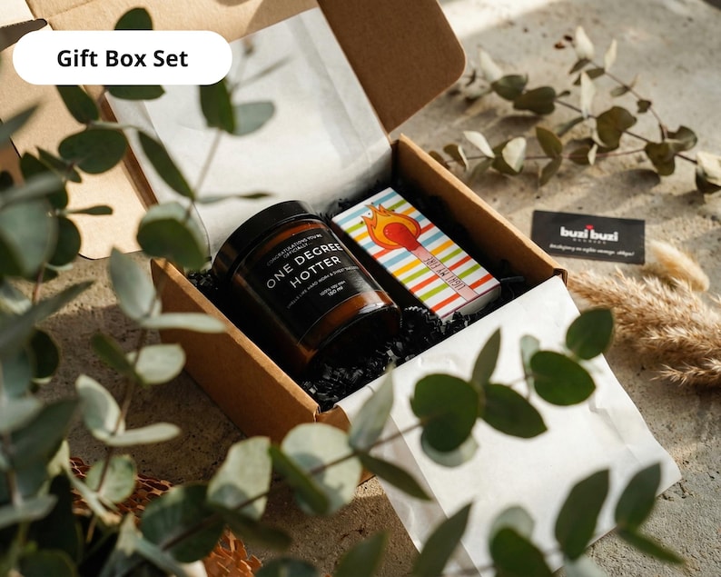 May include: A gift box set containing a jar of "ONE DEGREE HOTTER" candle, a matchbox with a colorful striped design, and a business card. The box is brown and lined with white paper and black filler.
