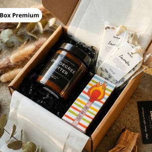 May include: A gift box with a candle, lavender soaps, and matches. The candle jar is brown with black text that reads "ONE DEGREE HOTTER". The matchbox has colorful stripes and the words "LIGHT MY FIRE". The box is filled with black crinkle paper.