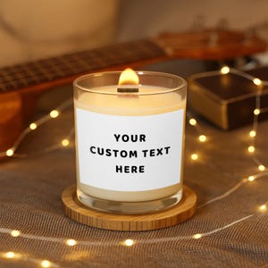 Può includere: Candela accesa in un barattolo di vetro trasparente con un'etichetta bianca che recita "YOUR CUSTOM TEXT HERE". La candela è appoggiata su un sottobicchiere rotondo di legno. La fiamma è accesa e lo sfondo include una chitarra e luci a corda.