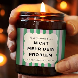 Könnte beinhalten: Eine brennende braune Glas-Kerze mit einem Etikett mit der Aufschrift "AB JETZT OFFIZIELL... NICHT MEHR DEIN PROBLEM AUF DEIN NÄCHSTES KAPITEL" in weißer Schrift auf grün-rosa gestreiftem Hintergrund. Die Kerze wird von einer Person in den Händen gehalten.