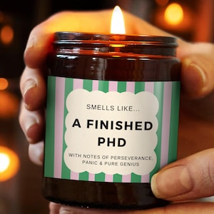 Può includere: Una candela accesa in un barattolo di vetro marrone con un'etichetta che recita "Smells Like... A Finished PHD" con testo sottostante. L'etichetta ha strisce verdi e rosa. La candela è tenuta tra le mani di una persona.