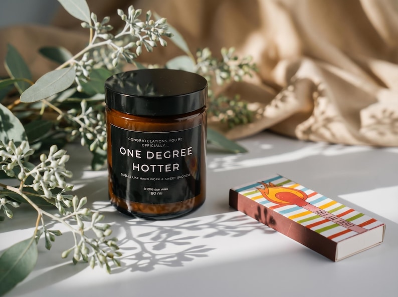 May include: A brown glass candle jar with a black lid and a label that reads "ONE DEGREE HOTTER." A colorful matchbox with a flame design sits beside the candle. Eucalyptus branches and a beige cloth are in the background.