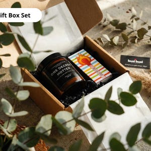 May include: A gift box set containing a jar labeled "ONE DEGREE HOTTER", a matchbox with a colorful striped design and a business card. The box is open, revealing the contents nestled on white tissue paper and black crinkle paper.