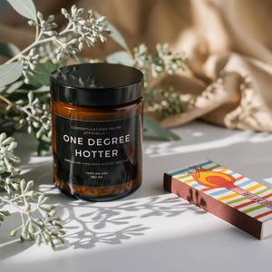 Puede incluir: Un tarro de vela de vidrio marr&oacute;n con tapa negra y una etiqueta que dice "ONE DEGREE HOTTER". Una caja de cerillas colorida con un dise&ntilde;o de llama se encuentra junto a la vela. Ramas de eucalipto y una tela beige en el fondo.