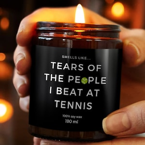 Puede incluir: Una vela encendida en un frasco de vidrio marrón. La etiqueta negra dice "Smells like... Tears of the people I beat at tennis." La vela es de 180 ml y está hecha de cera de soja 100%.