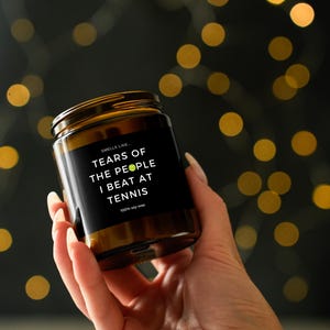 May include: A brown glass jar candle with a black label. The label reads "Smells like Tears of the people I beat at tennis." The candle is held in a hand against a blurred background of golden bokeh lights. The candle is made of 100% soy wax.