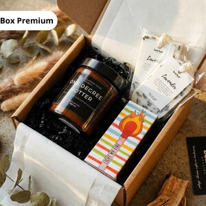 May include: A gift box with a candle, lavender soaps, and matches. The candle jar is brown with black text that reads "ONE DEGREE HOTTER". The matchbox has colorful stripes and the words "LIGHT MY FIRE". The box is filled with black crinkle paper.