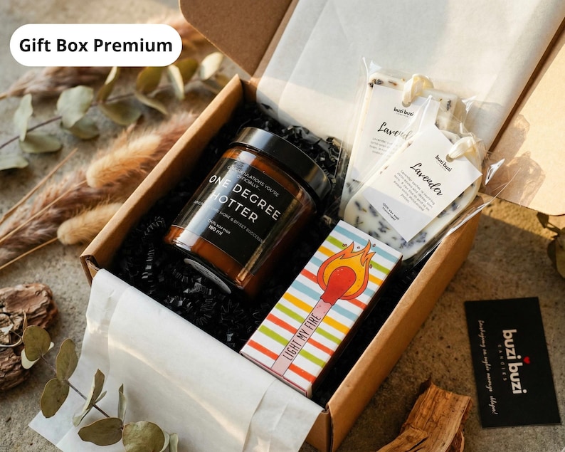 May include: A gift box with a candle, lavender soaps, and matches. The candle jar is brown with black text that reads "ONE DEGREE HOTTER". The matchbox has colorful stripes and the words "LIGHT MY FIRE". The box is filled with black crinkle paper.