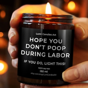 May include: A lit soy wax candle in a brown glass jar, held by a person. The black label reads "HOPE YOU DON'T POOP DURING LABOR. IF YOU DO, LIGHT THIS!" The candle is 180 ml.
