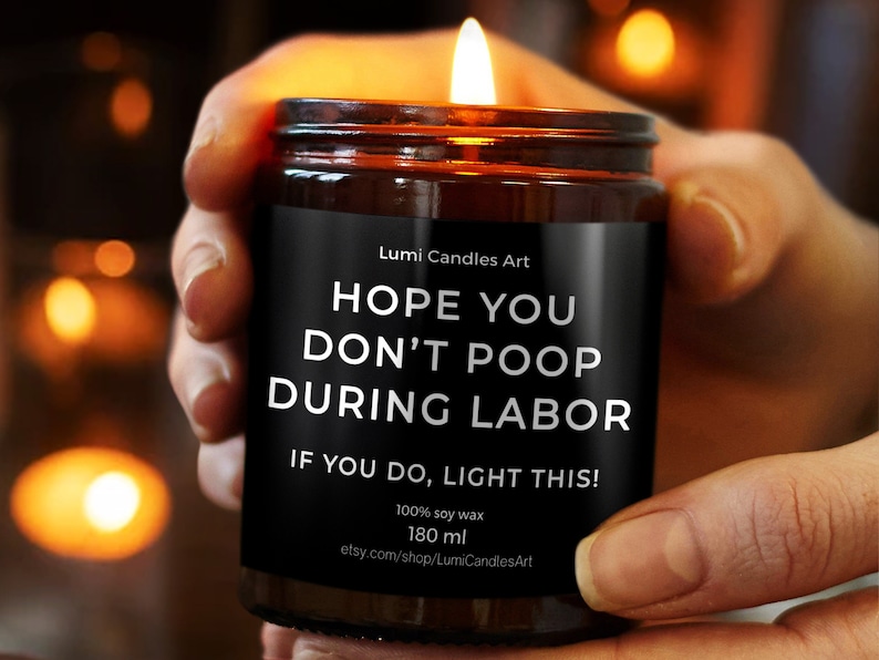 May include: A lit soy wax candle in a brown glass jar, held by a person. The black label reads "HOPE YOU DON'T POOP DURING LABOR. IF YOU DO, LIGHT THIS!" The candle is 180 ml.