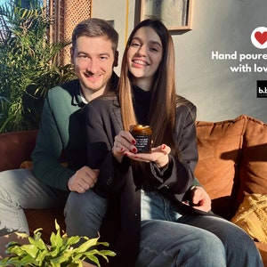 May include: A couple smiles while holding a small, black candle jar with a lit flame. The jar has white text on it. The words "Hand poured with love" and a heart icon are in the upper right corner. The candle is from b.b. candles.
