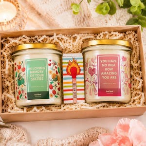 Op de afbeelding: Een geschenkdoos met twee glazen kaarsen en een klein luciferdoosje. De linkerkaars heeft een groen label met de tekst IN LOVING MEMORY OF YOUR SIXTIES. De rechterkaars heeft een roze label met YOU HAVE NO IDEA HOW AMAZING YOU ARE. Het luciferdoosje toont een illustratie van een lucifer en de tekst LIGHT MY FIRE.
