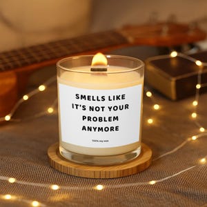Könnte beinhalten: Eine brennende Kerze in einem transparenten Glas mit einem Holzdocht, auf einem runden Holzuntersetzer. Die Kerze hat ein weißes Etikett mit dem Text "Smells Like It's Not Your Problem Anymore" in Schwarz. Unten steht "100% Soy Wax".