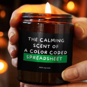 Op de afbeelding: Een bruine glazen kaars met een zwart etiket waarop "The Calming Scent of a Color Coded Spreadsheet" staat in witte tekst met een groene rechthoek om het woord "Spreadsheet". Het etiket vermeldt ook "100% soy wax".