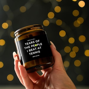 Puede incluir: Una vela en tarro de vidrio marr&oacute;n con una etiqueta negra. La etiqueta dice "Smells Like Tears of the People I Beat at Tennis". La vela se sostiene en una mano contra un fondo borroso de luces doradas bokeh. La vela est&aacute; hecha de cera de soja 100%.