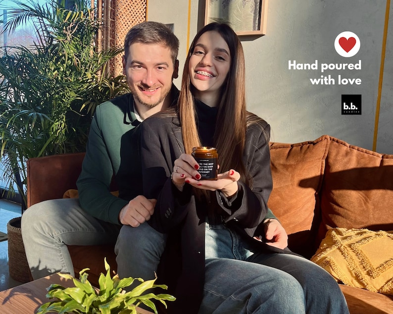 May include: A couple smiles while holding a small, black candle jar with a lit flame. The jar has white text on it. The words "Hand poured with love" and the brand logo are also visible.