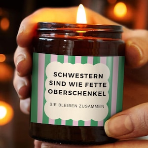 Könnte beinhalten: Eine brennende Kerze in einem braunen Glas. Das Etikett hat grüne und rosa Streifen und deutschen Text: "Schwester sind wie fette Oberschenkel, Sie bleiben zusammen." Die Kerze wird gehalten.