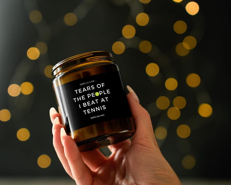 May include: A brown glass jar candle with a black label. The label reads "Smells like Tears of the people I beat at tennis." The candle is held in a hand against a blurred background of golden bokeh lights. The candle is made of 100% soy wax.