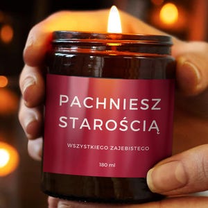 May include: A lit candle in a brown glass jar with a red label. The label has white text that reads "PACHNIESZ STAROŚCIĄ" and "WSZYSTKIEGO ZAJEBISTEGO". The jar contains 180 ml of wax.