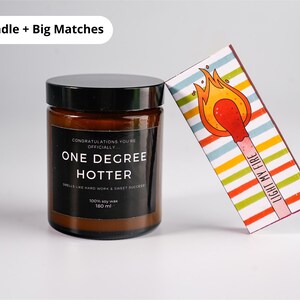 May include: A brown glass jar candle with a black lid and a label that reads "ONE DEGREE HOTTER." A decorative matchbox with a colorful striped design and a lit match illustration is leaning against the candle. The text "LIGHT MY FIRE" is printed on the matchbox.
