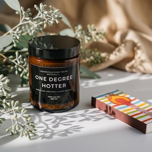 May include: A brown glass candle jar with a black lid and a label that reads "ONE DEGREE HOTTER." A colorful matchbox with a flame design sits beside the candle. Eucalyptus branches are in the background.