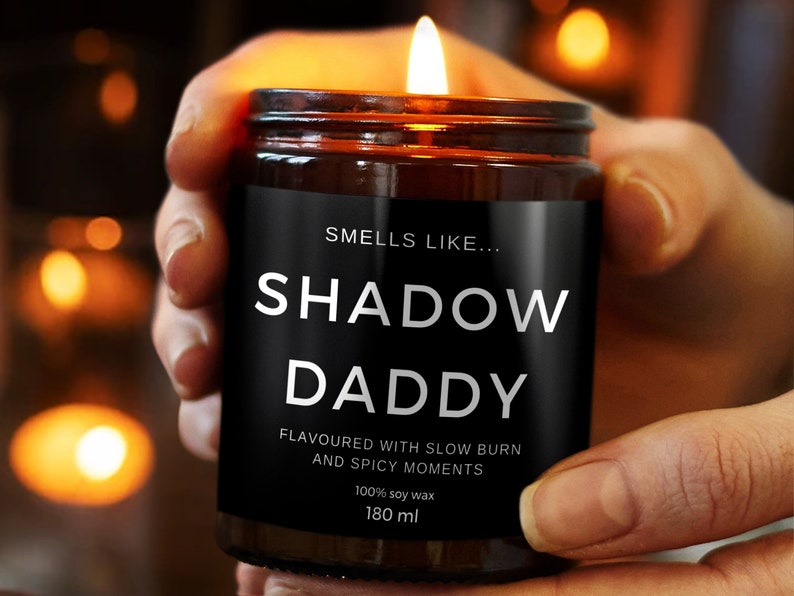May include: A lit candle in a brown glass jar held by a person. The black label reads "SMELLS LIKE... SHADOW DADDY" with additional text below. The candle is made of 100% soy wax and is 180 ml.