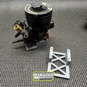 Puede incluir: Un soporte de motor gris impreso en 3D con un motor negro y plateado adjunto. El motor tiene una etiqueta roja y plateada que dice "M Line". El soporte del motor tiene un diseño triangular y está etiquetado como "WEBSTER MODS".