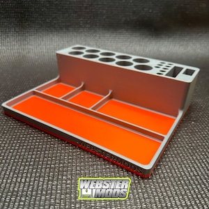 May include: A grey 3D printed organiser with a bright orange tray. The organiser has multiple compartments for storing small items. The tray has the text "WEBSTERMODS" printed on it.