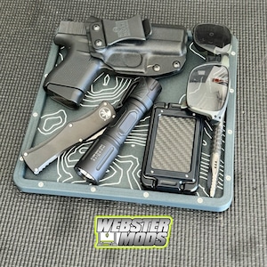 May include: A collection of everyday carry items arranged on a gray tray with a white topographic design. Items include a black handgun in a holster, a flashlight, a knife, sunglasses, a phone case, and a pen. The tray has the text "WEBSTER MODS".