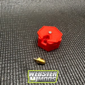 以下が含まれることがあります： 中央に小さな穴があり、金色の金属製コネクターが付いた赤い3Dプリント六角形ツール。このツールは3Dプリントを支援するために設計されています。