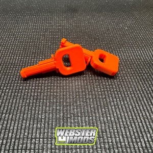 May include: Two bright orange plastic keys, one with a longer handle, are lying on a black textured surface. The keys are designed to look like a traditional key with a keyhole and a handle.