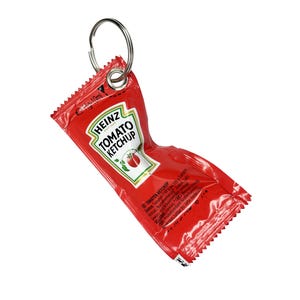 Puede incluir: Un sobre de ketchup Heinz rojo con un llavero plateado adjunto. El sobre presenta el logotipo de Heinz y una ilustración de tomate. El texto "TOMATO KETCHUP" es visible. El sobre está arrugado.