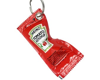 Könnte beinhalten: Ein rotes Heinz Tomaten Ketchup-Päckchen mit einem silbernen Schlüsselring. Das Päckchen zeigt das Heinz-Logo und eine Tomaten-Illustration. Der Text "TOMATO KETCHUP" ist sichtbar. Das Päckchen ist zerknittert.